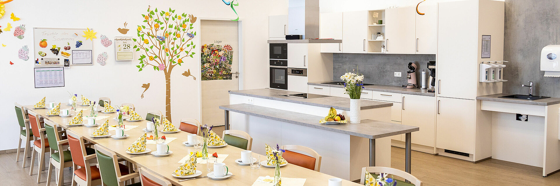 BRK-Tagespflegen in Eggenfelden und Gangkofen Als Beispielbild für die BRK-Tagespflegen in Eggenfelden und Gangkofen dient eine Innenansicht des Aufenthaltsraumes in Eggenfelden. Ein langer Tisch ist sommerlich gedeckt. Auf den weißen Tellern sind Servietten mit Zitronenmuster zu sehen. Im Hintergrund ist die Gruppenküche zu sehen.
