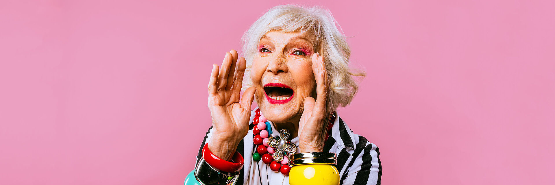 Kostenloser Schnuppertag bei den BRK-Tagespflegen Rottal-Inn Eine ältere Dame mit weißem Haar nimmt die Hände zum offenen Mund um freudig etwas anzukündigen. Die Frau ist grell geschminkt und trägt bunte große Perlenketten und Armreifen.